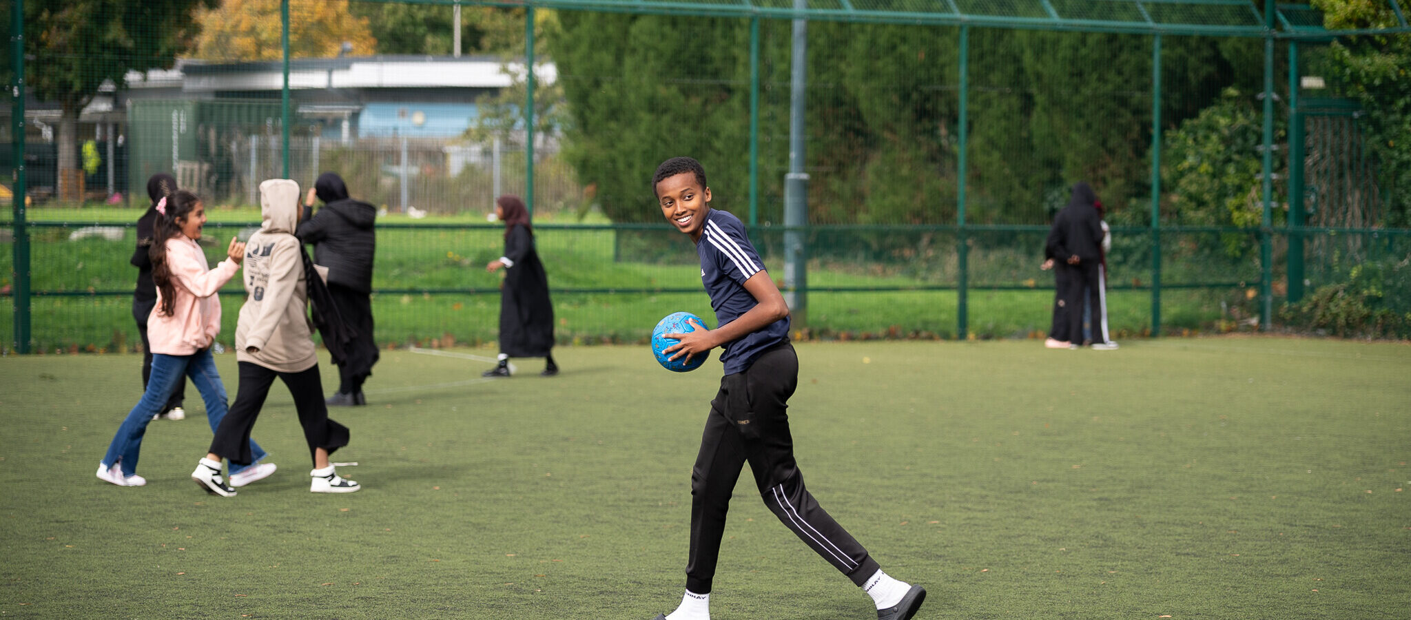 A young person holding a blue football on a pitch, there are others in the background.