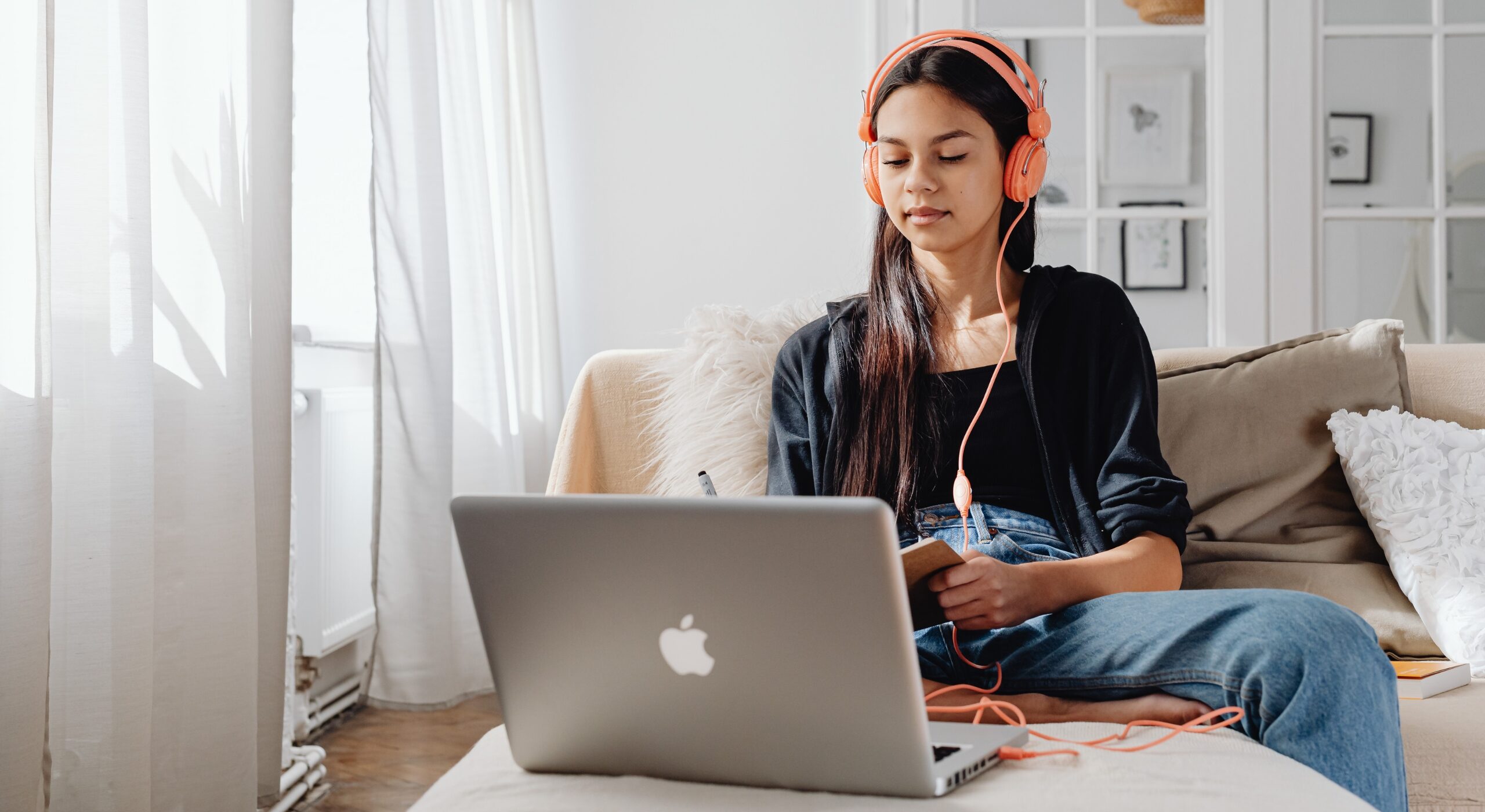 Young person sitting on a sofa at home using a laptop while wearing headphones and holding a notebook. The image represents online learning, digital skills development, and flexible study from home.