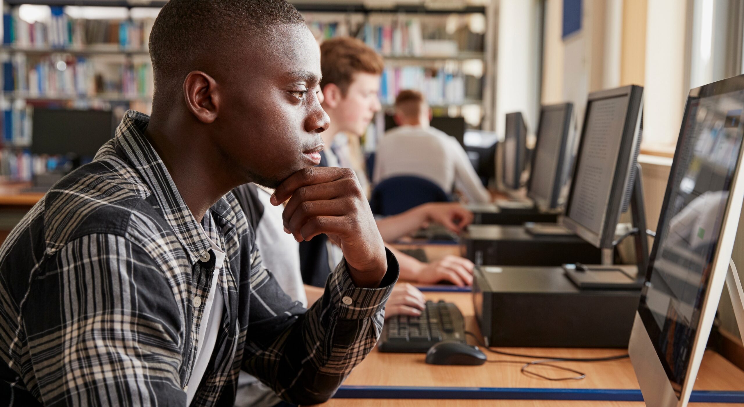 Young person using a desktop computer in a library or study environment. The person is seated at a desk, resting their chin on one hand while looking at the computer screen. Several other people are working at computers in the background, with bookshelves visible behind them. The image is used to promote IBM SkillsBuild on the DofE website and highlights digital learning, skills development and online education opportunities.