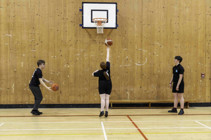 A basketball practice scene in a gymnasium with a wooden wall. A girl with red hair is shooting a basketball towards the hoop, while a boy in glasses stands nearby, holding a basketball and preparing to pass. Another boy watches from the side, both wearing black athletic shirts. The gym floor is marked with lines for basketball.