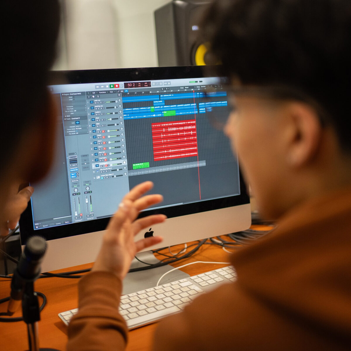 Person using audio editing software on a desktop computer, with a microphone and keyboard visible in the foreground. The screen displays multiple audio tracks being edited, representing digital media creation, content production and creative skills development.