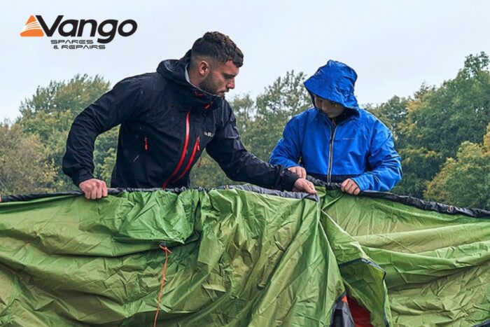 Two people in waterproof jackets assemble a large green tent outdoors in wet weather, with trees in the background. The Vango Spares and Repairs logo is in the corner.