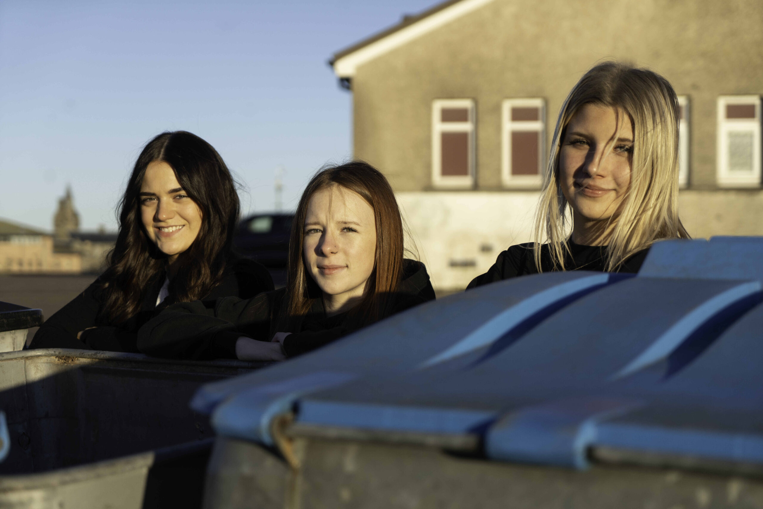 Three young women are leaning against blue and gray dumpsters in an urban setting. They are smiling and looking at the camera, with a clear blue sky in the background. The buildings behind them suggest a city environment. The women have varying hair colors and styles, and they are dressed in casual black clothing.