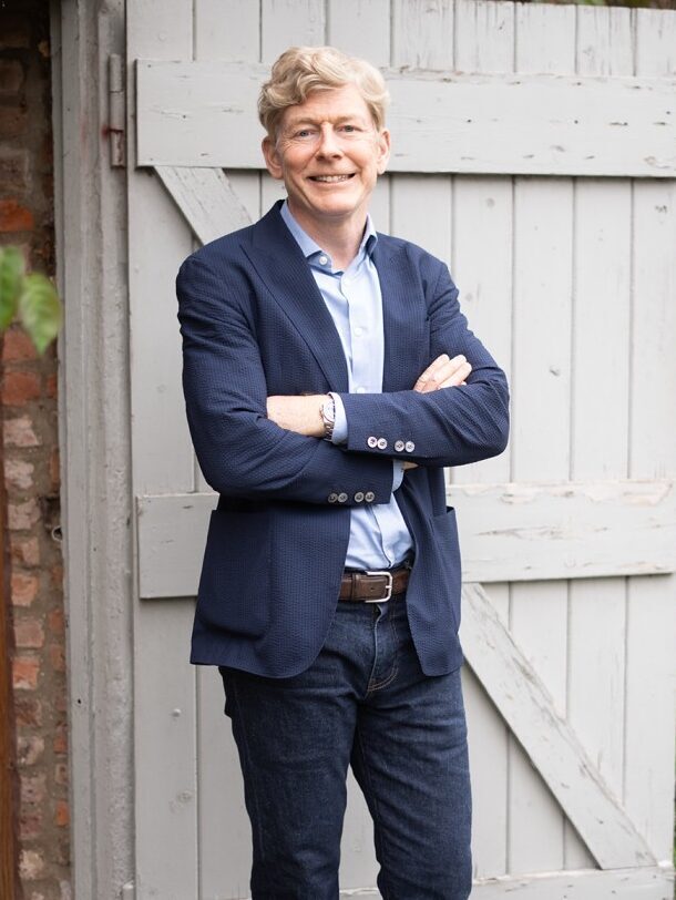 Richard stands in front of a white door wearing a dark blue suit.