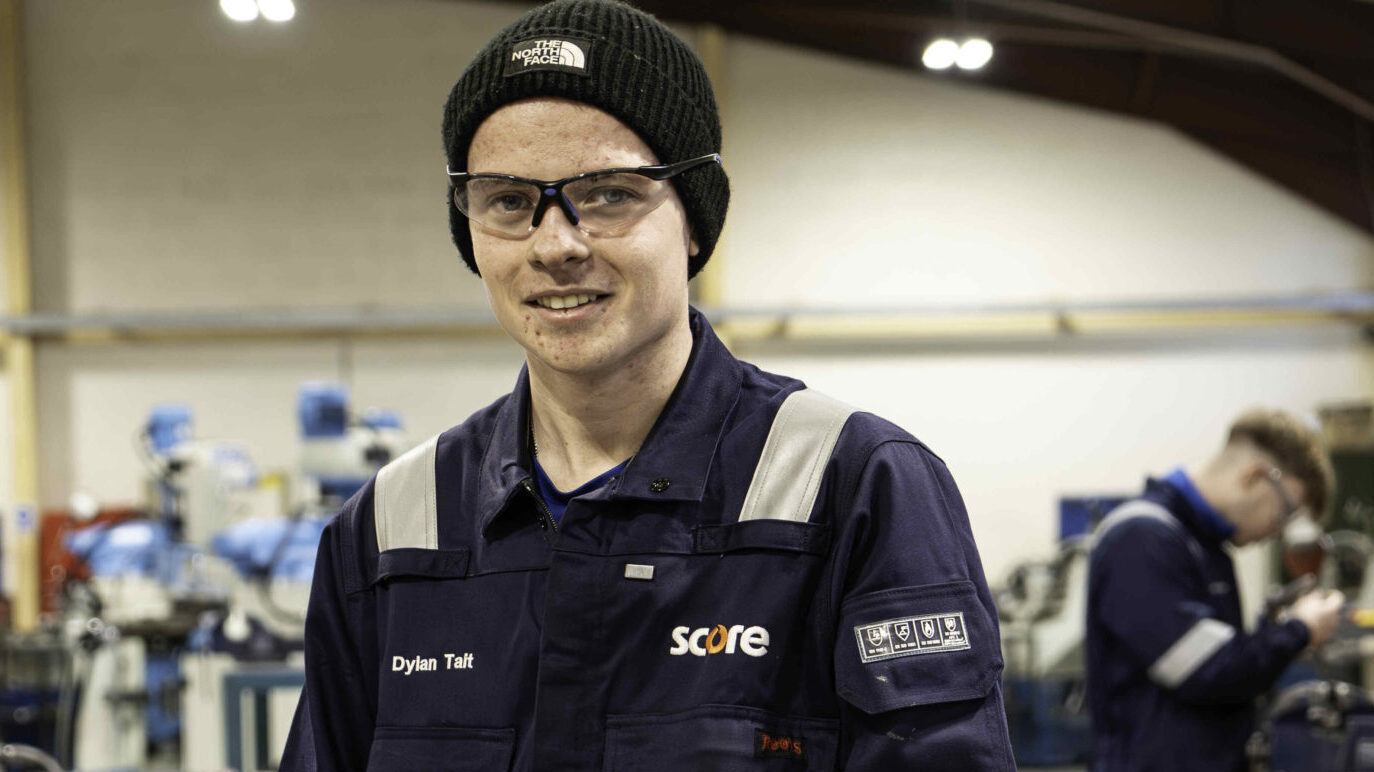 Dylan in safety overalls holding a hacksaw in a workshop, with workbenches and tools in the background.