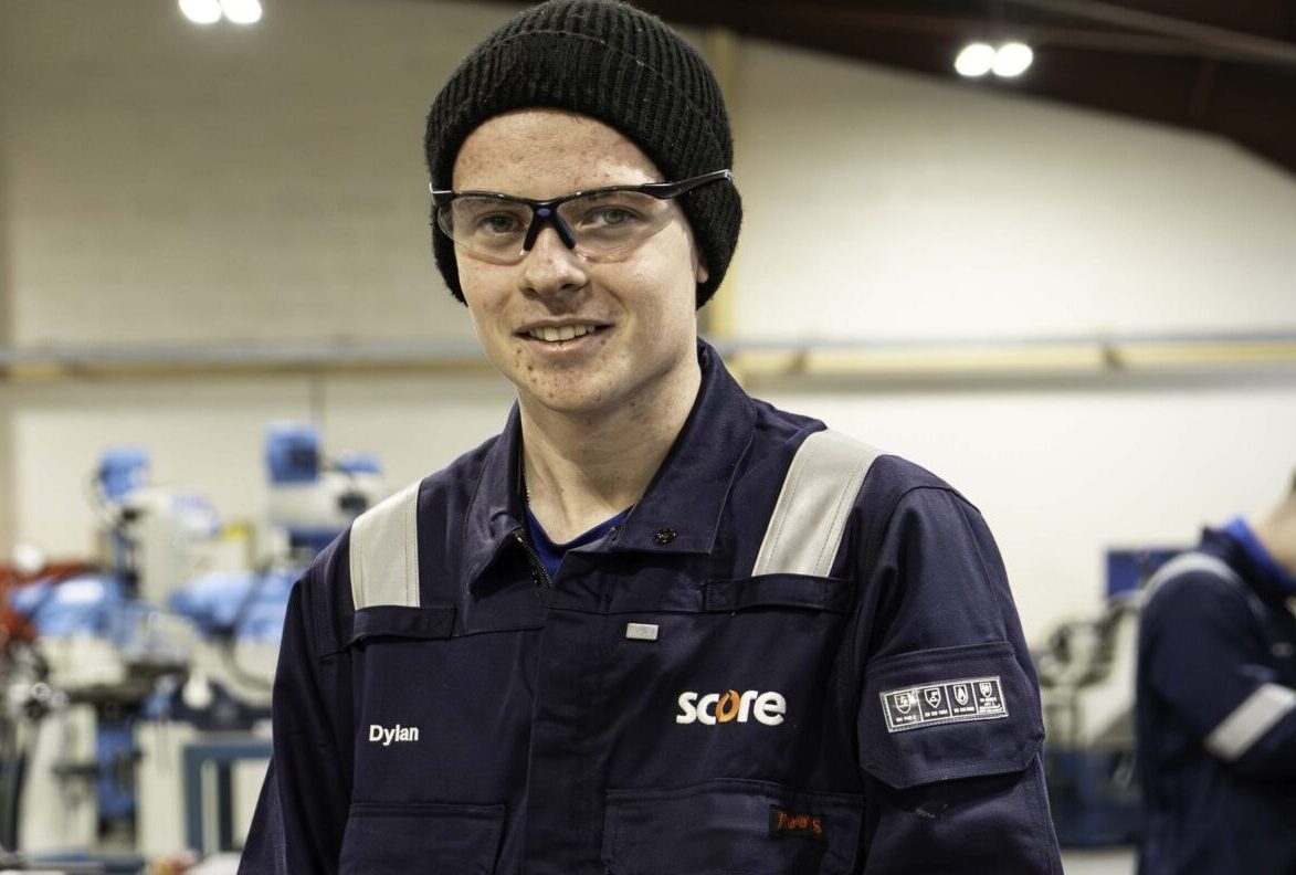 Dylan in safety overalls holding a hacksaw in a workshop, with workbenches and tools in the background.