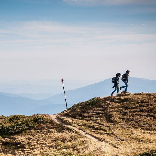 two young people walk in nautre on large hill with mountains in background