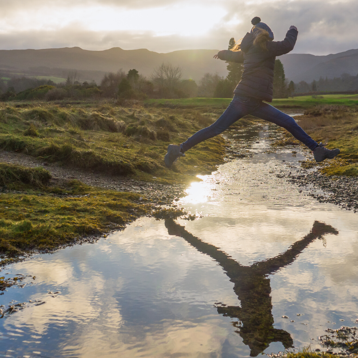 Girl in jacket and woolly hat jumping over stream