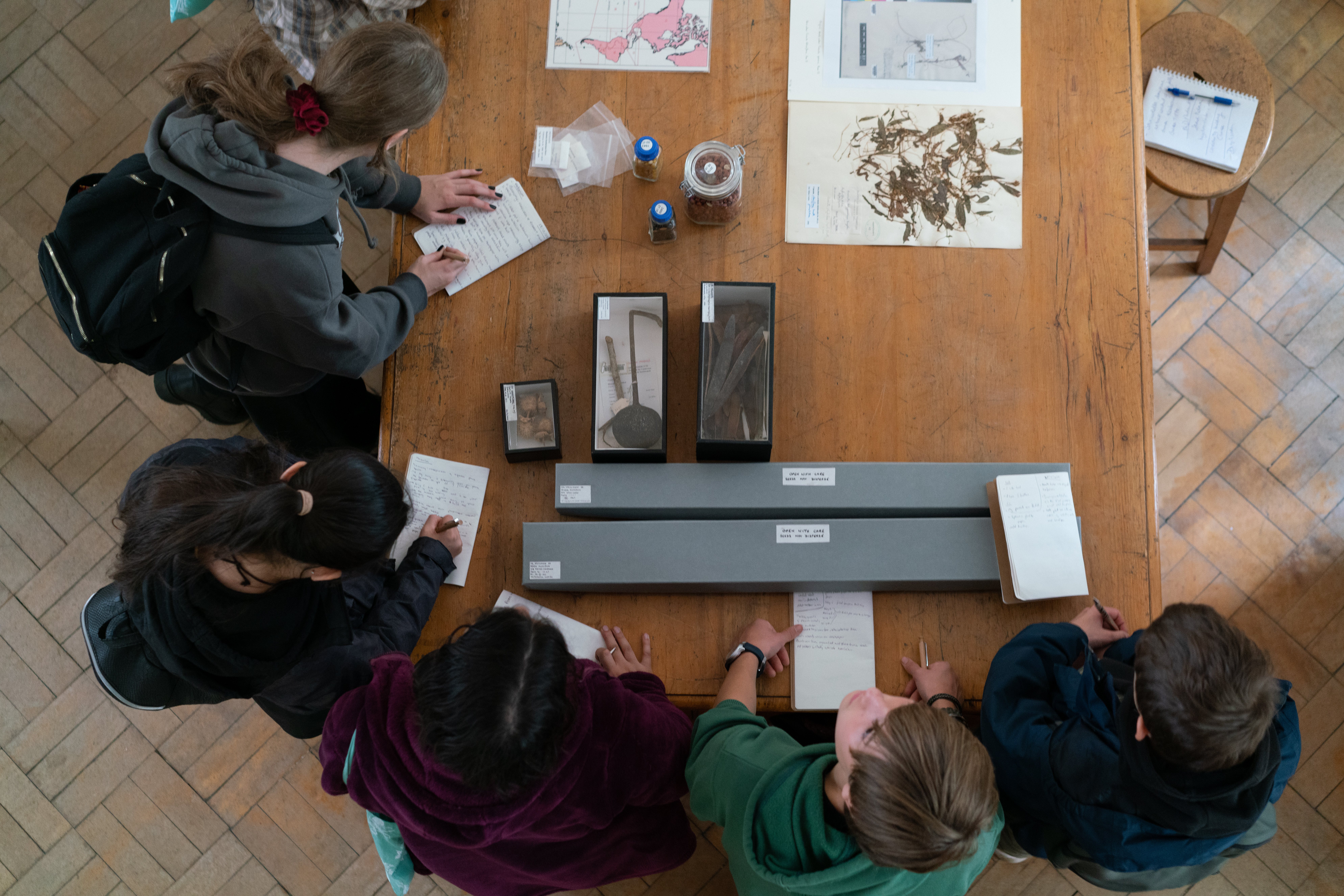 Young people sitting around a large table with boxes and objects on it writing notes.