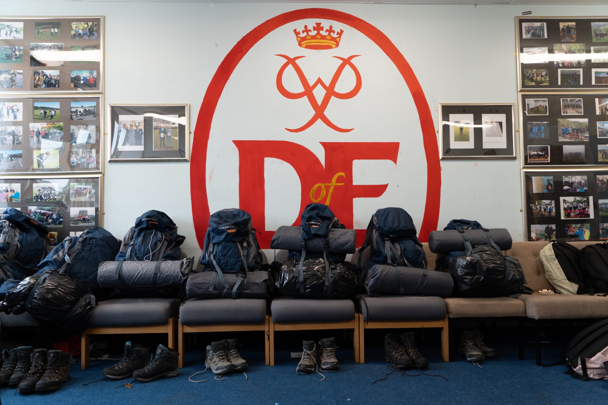 Rucksacks sitting on chairs in front of a large DofE logo painted on the wall.