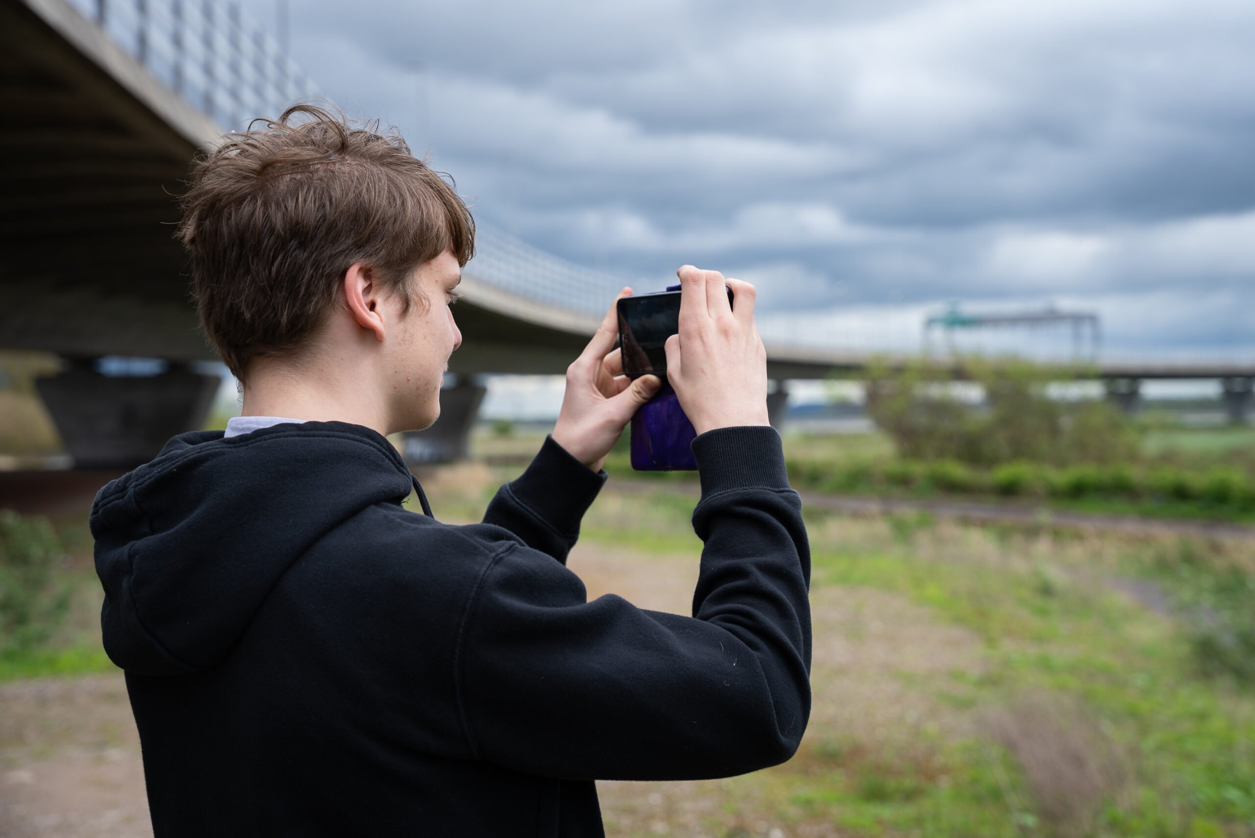 A young person taking a picture of grasslands and a bridge using their phone.
