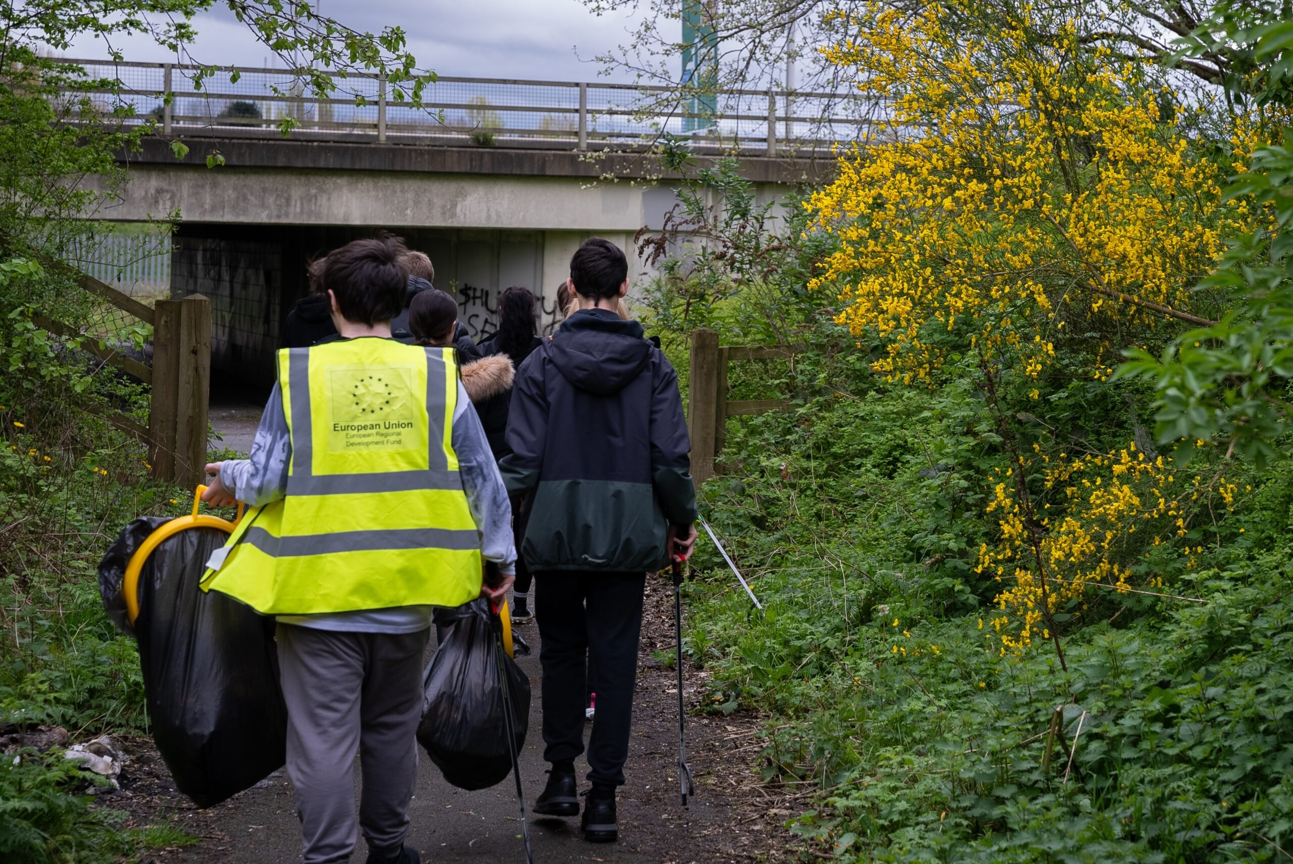 A group of young people, one of them wearing high-vis, doing a litter pick along a path with greenery.