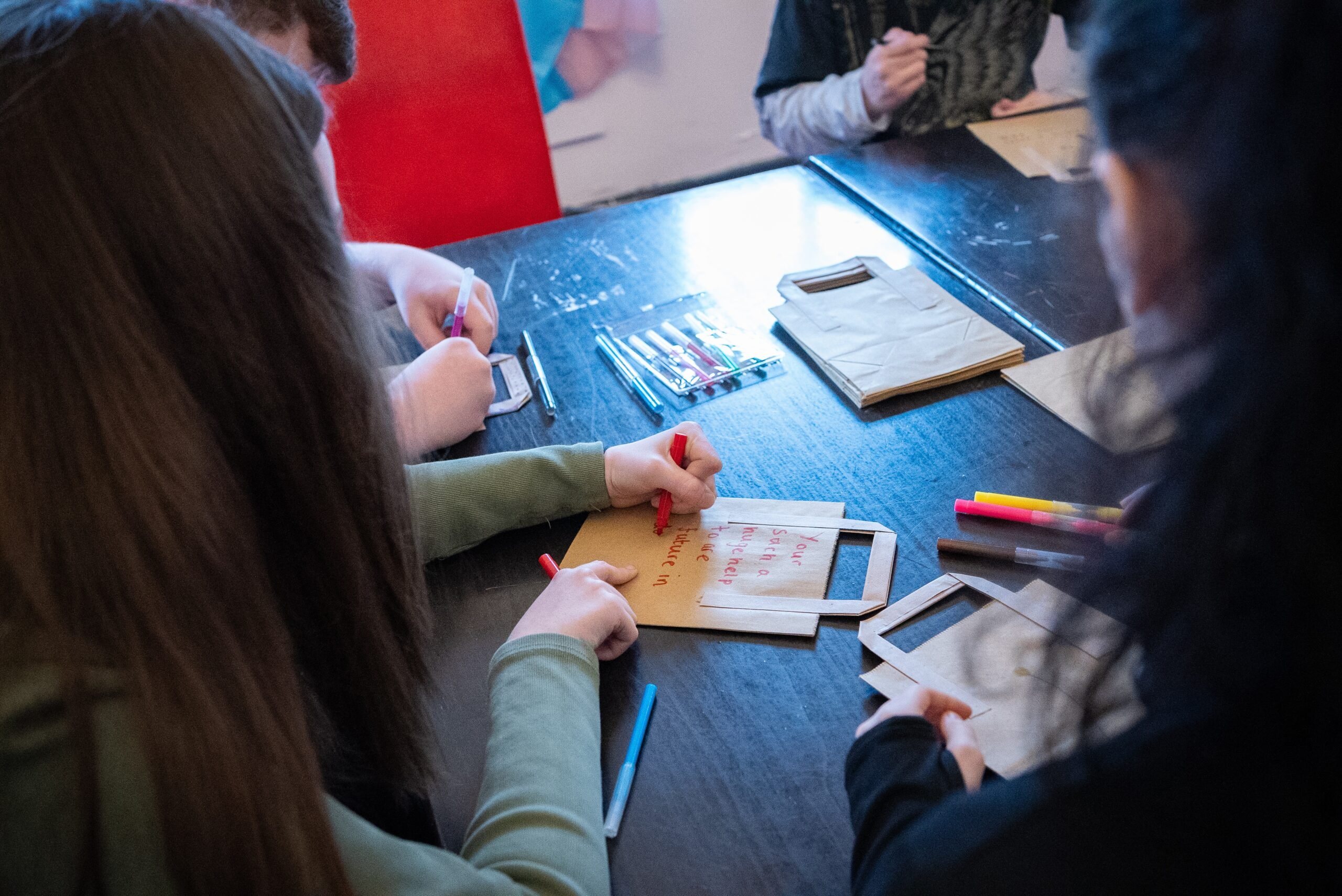 A young person sitting at a table writing on a brown paper bag with a red pen.