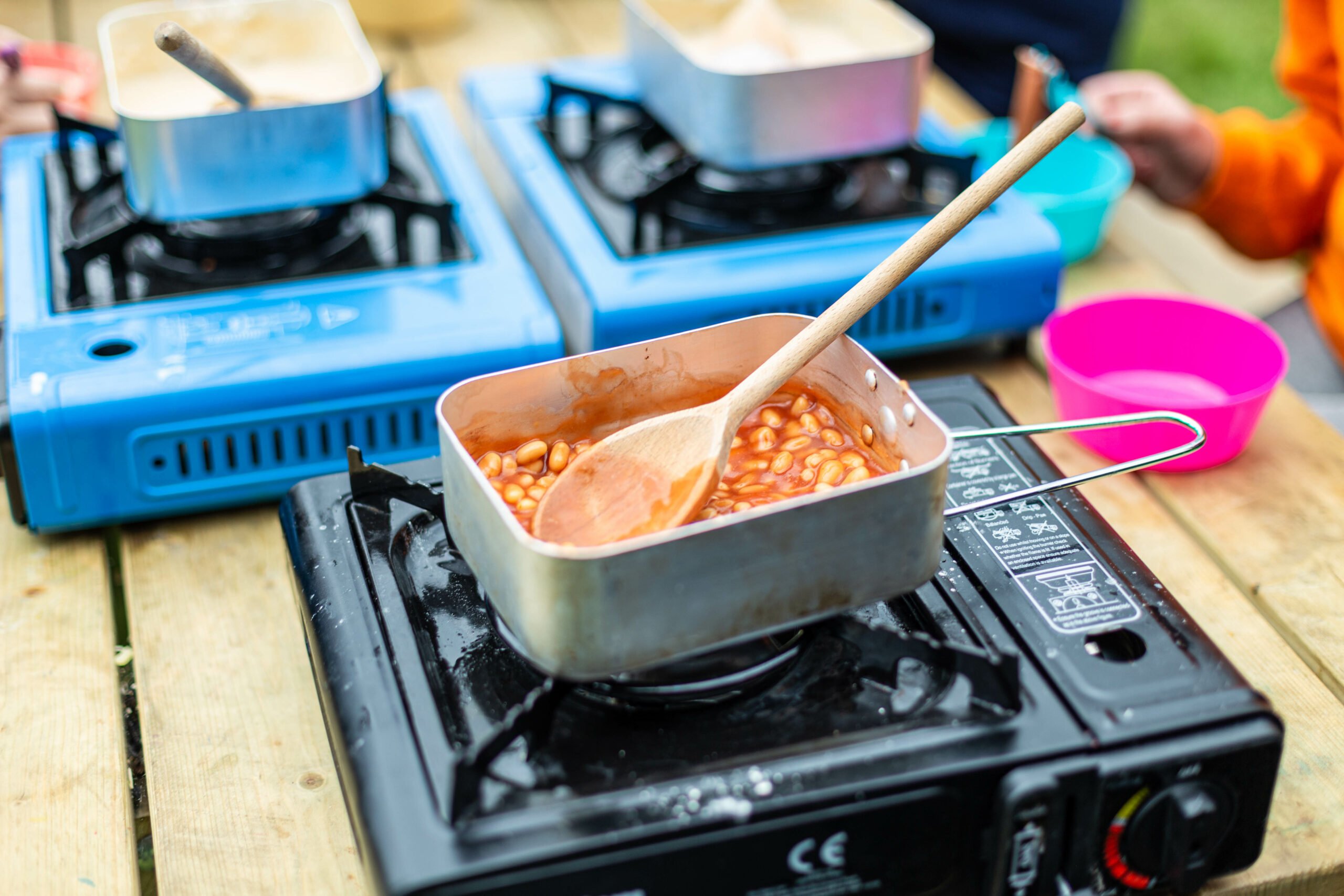 Baked beans and a wooden spoon in a metal box on an outdoors gas cooker.