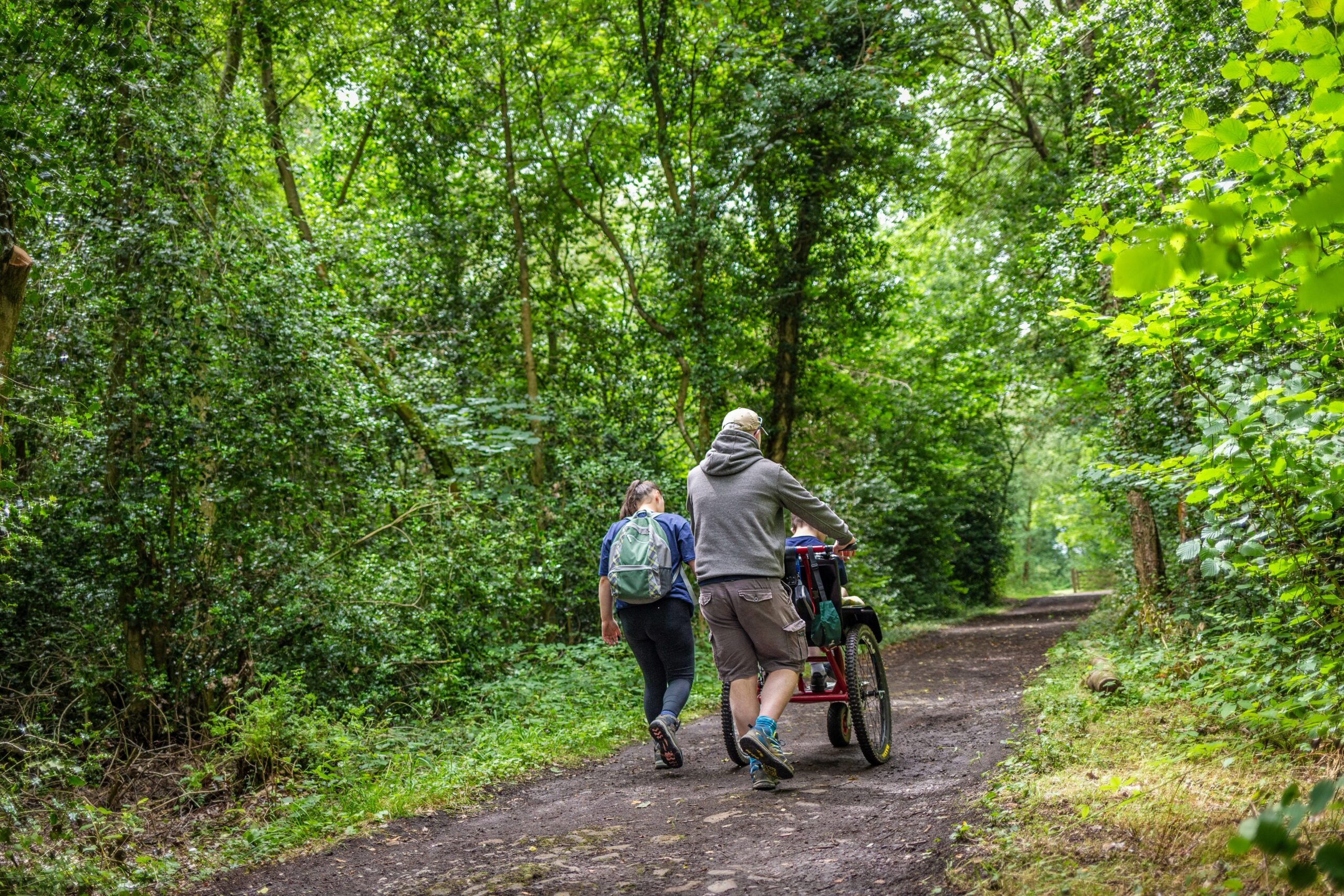 Three people on a woodland path, one is walking, and one is pushing a wheelchair the third person sits in.