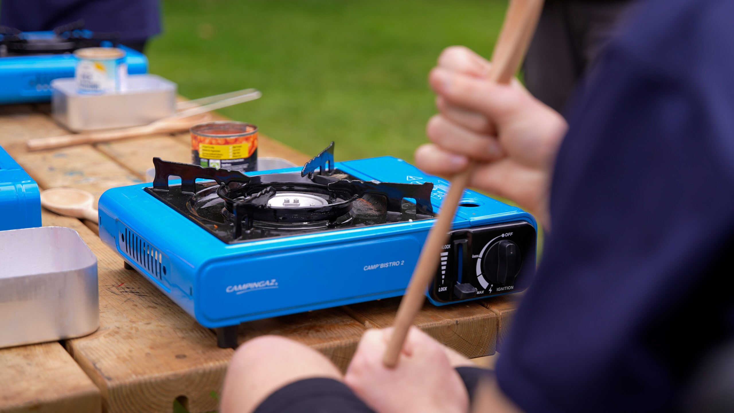 A blue camping gas stove on a wooden table, a young person holding a wooden spoon is sitting next to it.