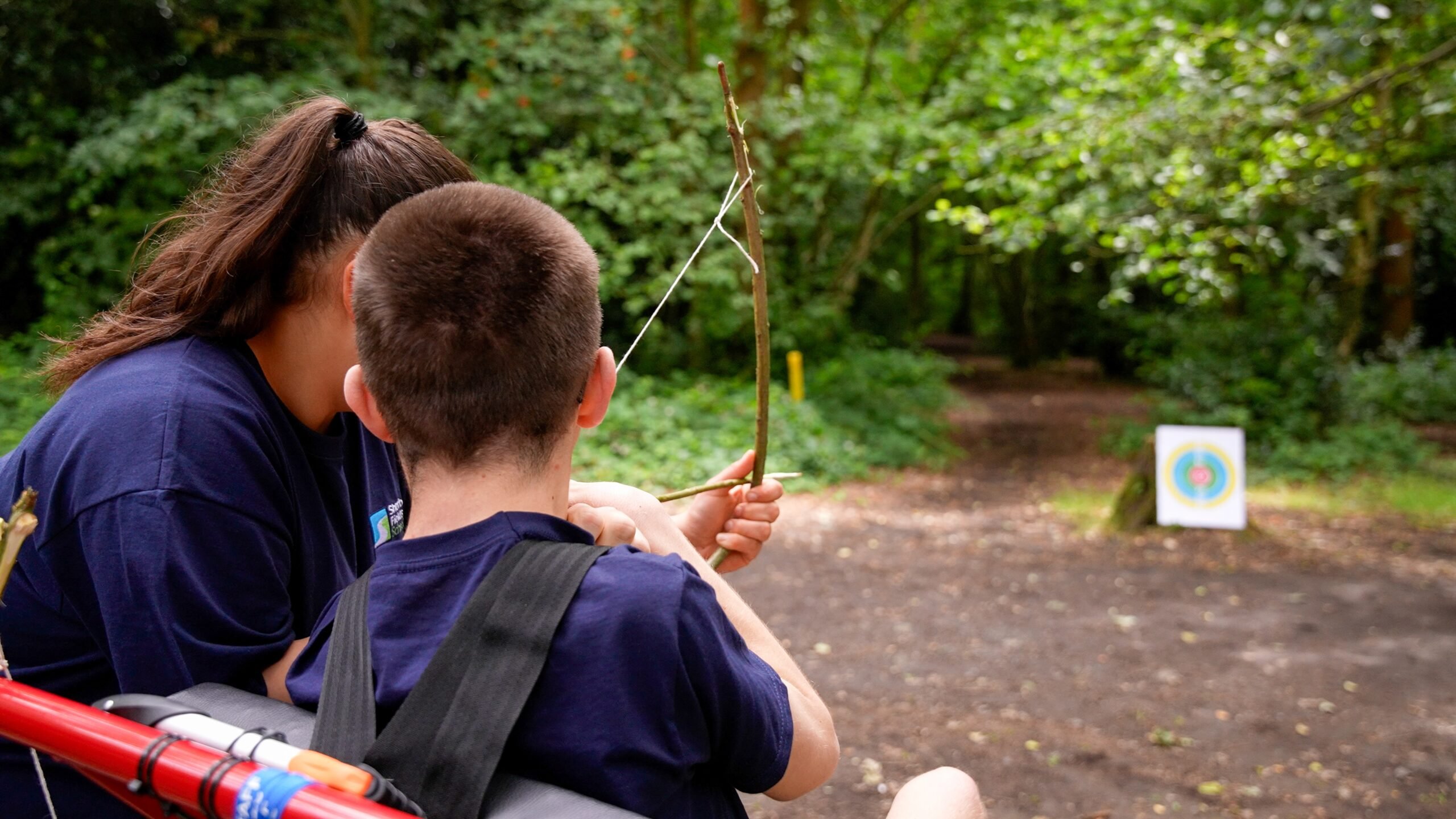 A young person in a wheelchair being shown how to aim at a target using a bow and arrow.