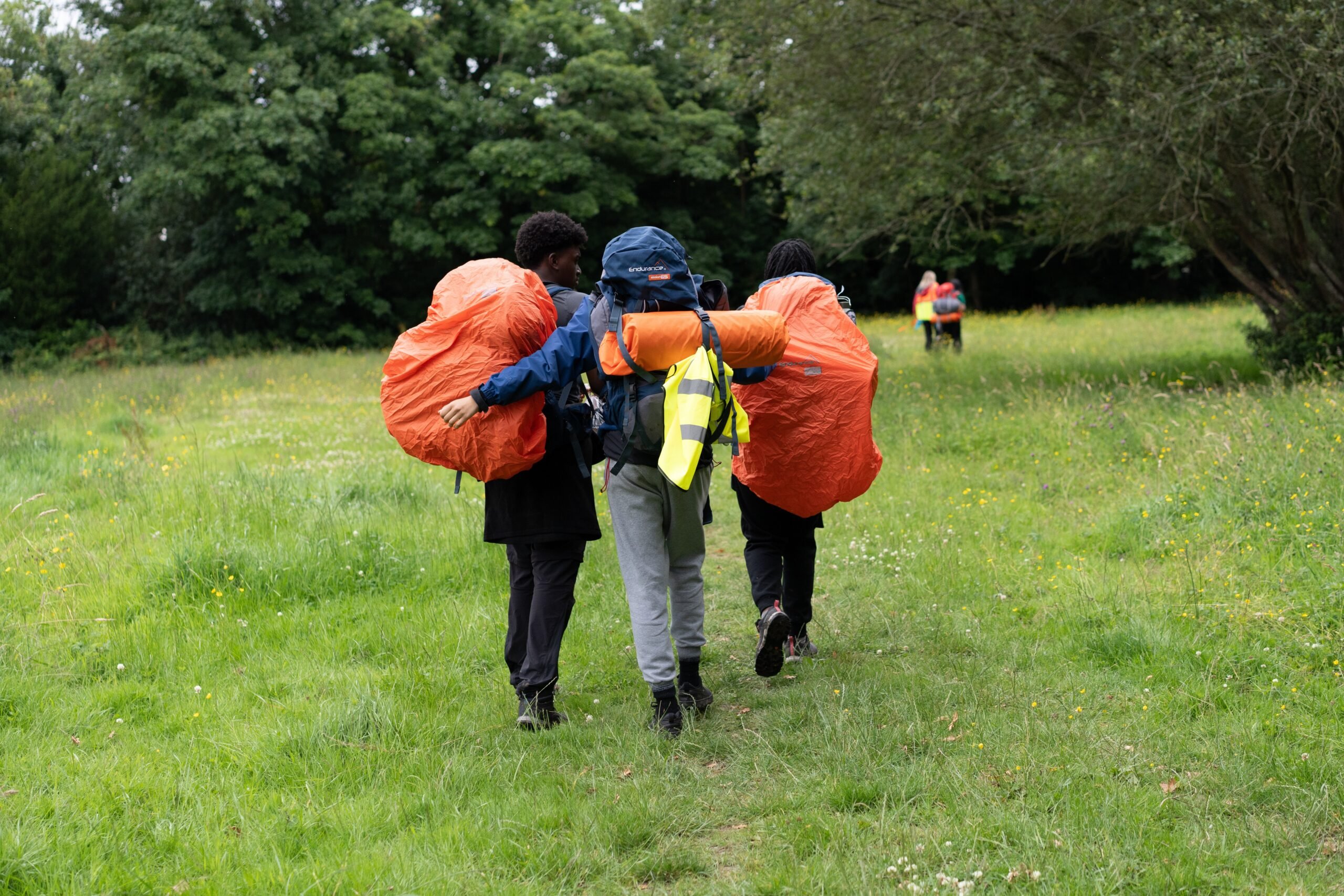 Three young people with full rucksacks walking together across a grassy meadow.