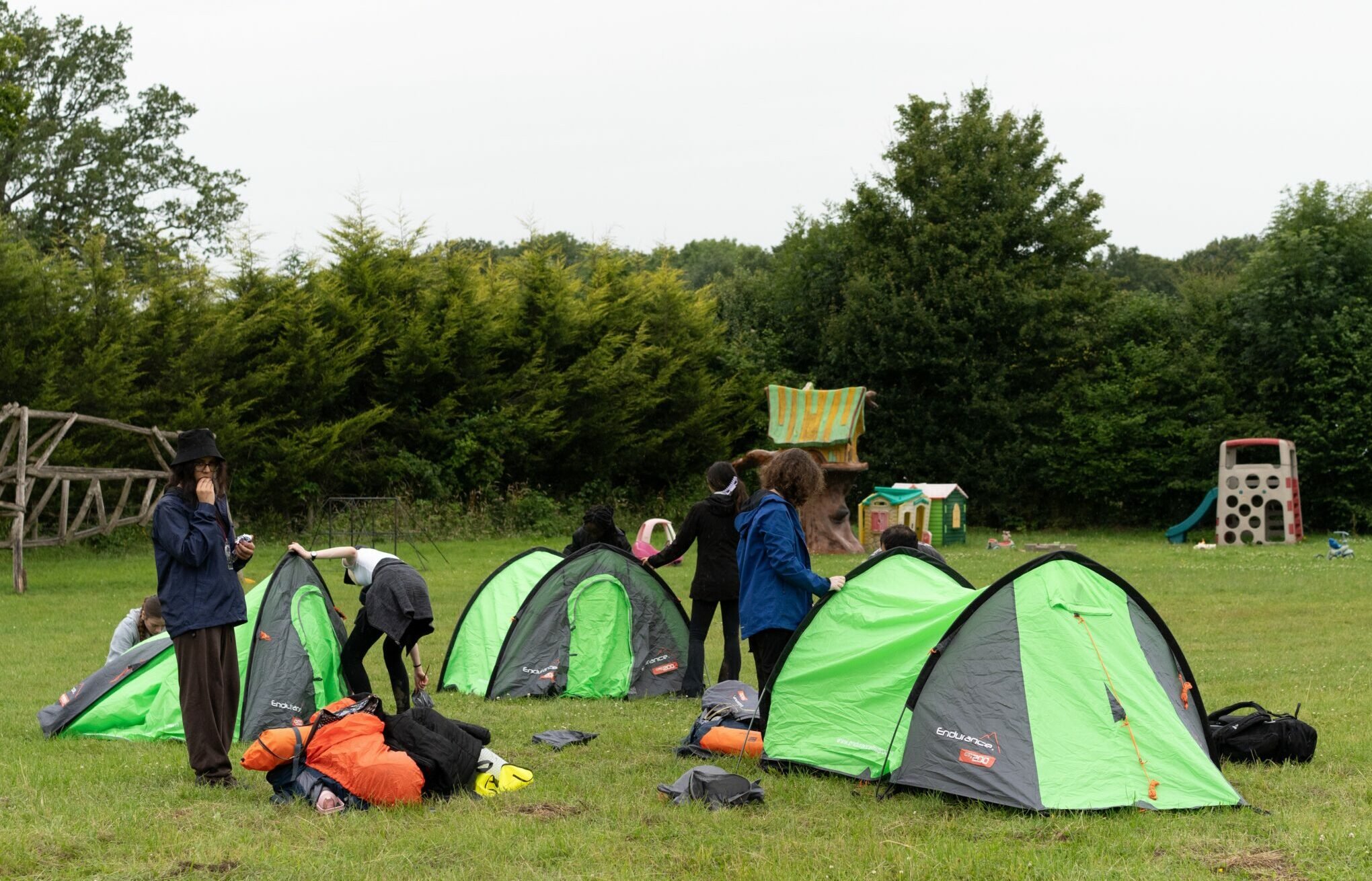 Young people putting up three tents on a campsite surrounded by trees.