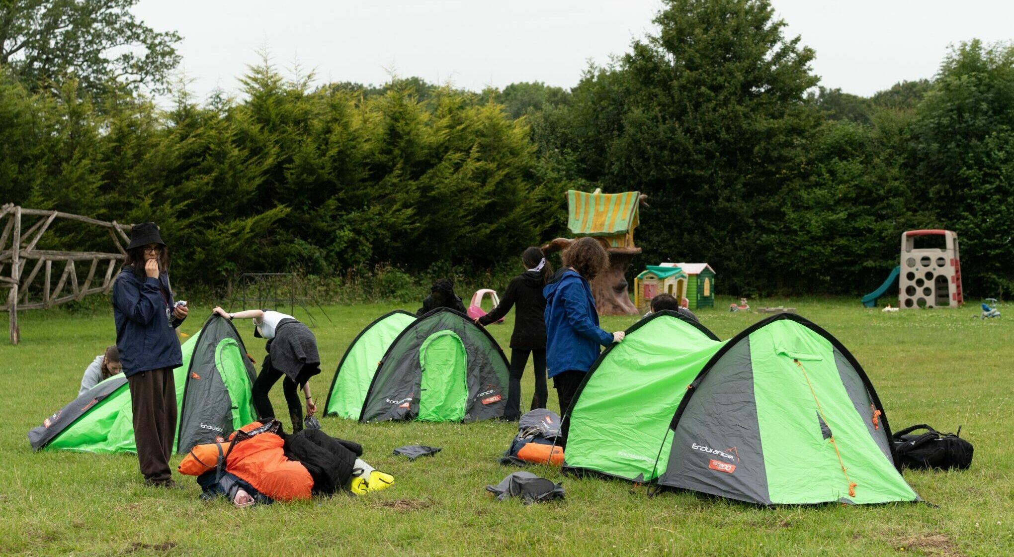 Young people putting up three tents on a campsite surrounded by trees.