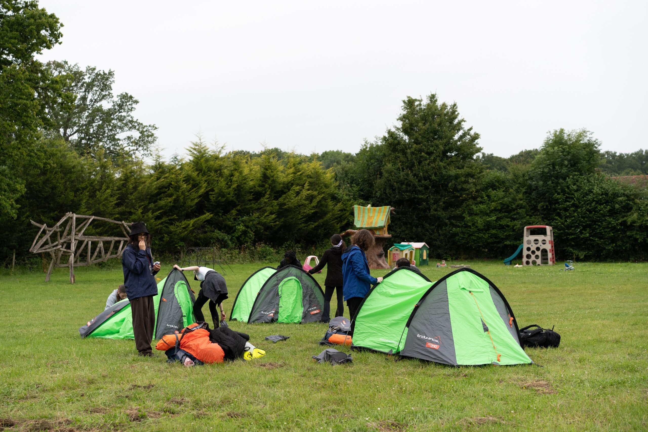 Young people putting up three tents on a campsite surrounded by trees.