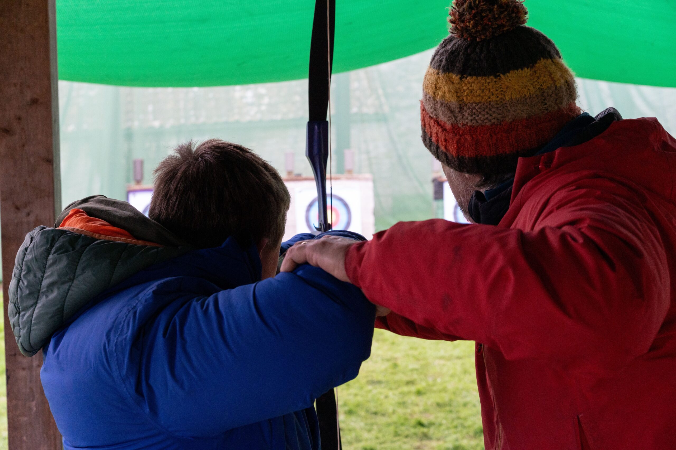 A volunteer helping a young person aim at a target with a bow and arrow.