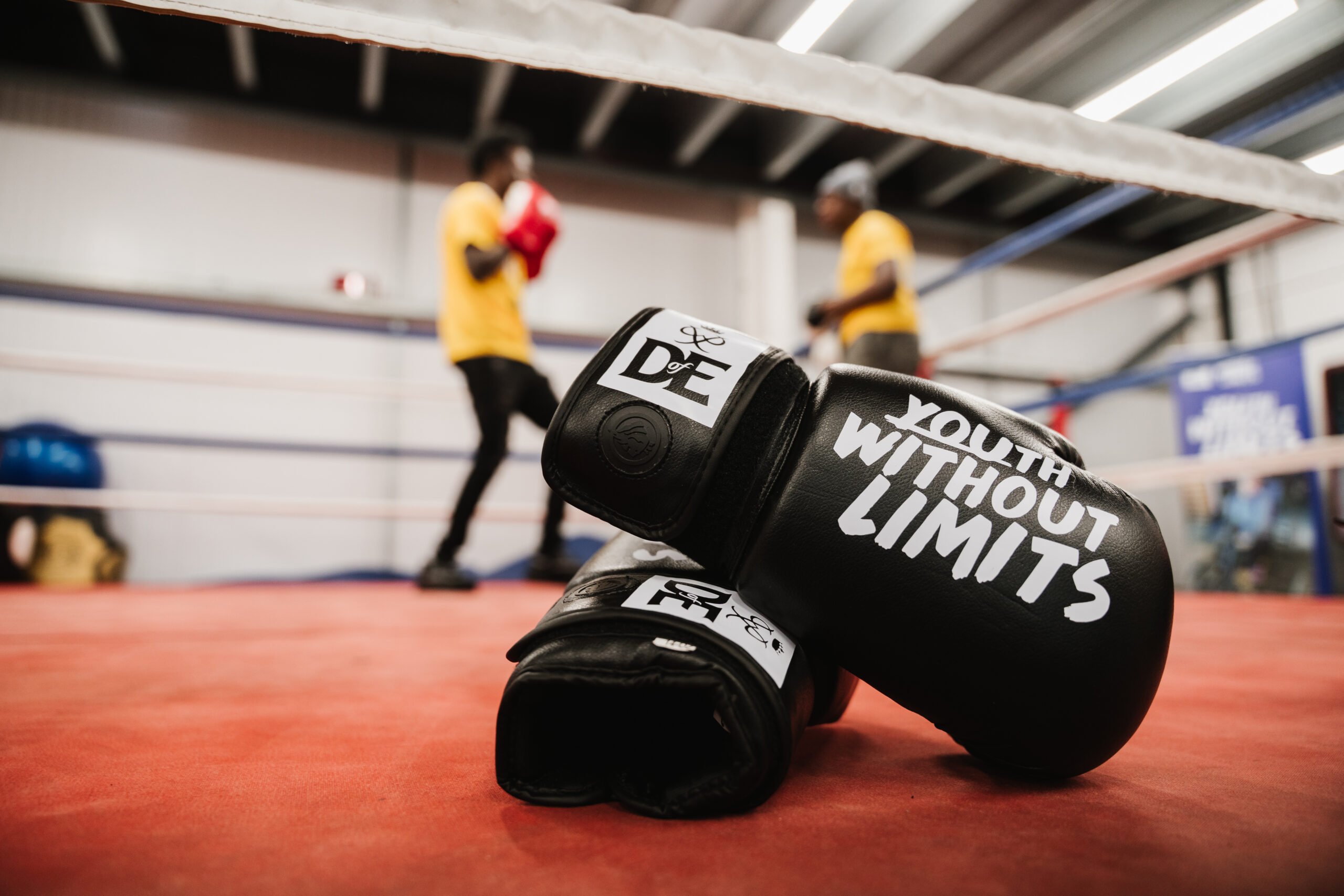 A pair of black boxing gloves saying Youth Without Limits lying in a boxing ring with two people in it.