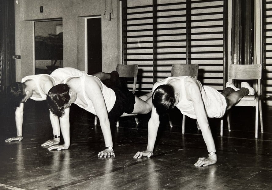 Three individuals are performing a physical exercise in a gymnasium, positioned in a straight line on a polished wooden floor. Each person is in a plank-like stance with arms extended and hands flat on the floor, while their legs are elevated and supported by a wooden gym bench behind them. They are wearing light-colored sleeveless tops and dark shorts. In the background, there are wooden wall bars and a door with a window panel.